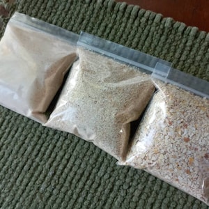 May include: Three clear plastic bags filled with different types of granular substances. The bags are sealed with a zip closure. The contents vary in color, ranging from light beige to a mix of beige and brown. The bags are arranged on a textured green surface.