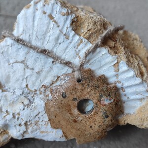 May include: A brown ceramic pendant with a circular hole in the center, hanging from a beige cord. The pendant is resting on a white and brown rock.