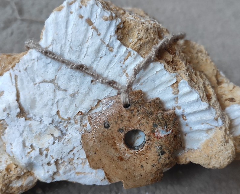 May include: A brown ceramic pendant with a circular hole in the center, hanging from a beige cord. The pendant is resting on a white and brown rock.