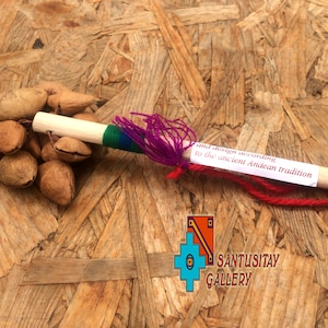 May include: A wooden stick with a purple and green tassel attached to one end. A cluster of brown seed pods are attached to the other end. The stick has a white label with the text "and design according to the ancient Andean tradition". The stick is on a wooden surface. The text "SANTUSITAY GALLERY" is visible in the lower right corner of the image.