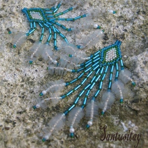 Op de afbeelding: Een paar hangende oorbellen met blauwe en groene kralen en witte plastic accenten. De oorbellen hangen aan een ruw, grijs oppervlak.