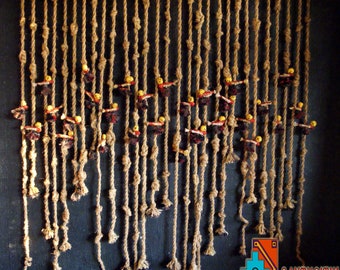 May include: A wall hanging made of brown rope with small, colorful figures woven into the strands. The figures are arranged in rows and appear to be wearing yellow hats. The hanging is against a dark background.