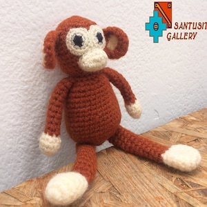 May include: A brown crocheted monkey toy with white accents on the ears, hands, and feet. The monkey is sitting on a wooden surface.