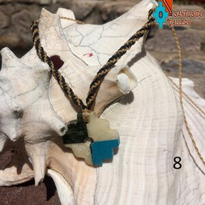 May include: A turquoise, green, and white stone pendant in the shape of a stylized heart, hanging from a black and gold braided cord. The pendant is resting on a white seashell. The text "SANTUSITAY GALLERY" is visible in the upper right corner of the image.