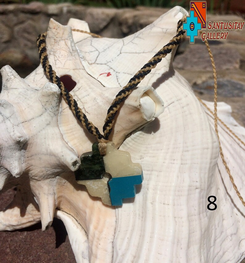 May include: A turquoise, green, and white stone pendant in the shape of a stylized heart, hanging from a black and gold braided cord. The pendant is resting on a white seashell. The text "SANTUSITAY GALLERY" is visible in the upper right corner of the image.