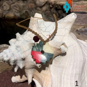 May include: A red, white, green, and black stone pendant with a round hole in the center, hanging from a brown cord. The pendant is on a white seashell.