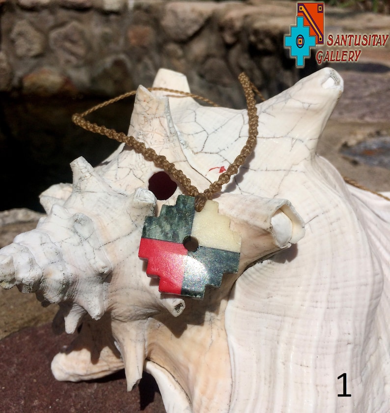 May include: A red, white, green, and black stone pendant with a round hole in the center, hanging from a brown cord. The pendant is on a white seashell.