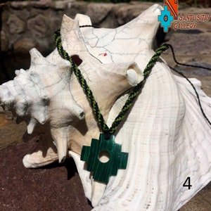 May include: A green jade stone pendant in the shape of a cross with a black and green braided cord. The pendant is sitting on a white seashell.