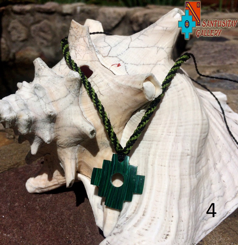 May include: A green jade stone pendant in the shape of a cross with a black and green braided cord. The pendant is sitting on a white seashell.
