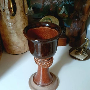 May include: A brown ceramic chalice with a wide, open top and a pedestal base. The chalice has a glossy finish and a decorative ring around the stem. The base is a lighter shade of brown.