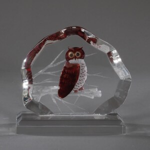 May include: A clear glass owl figurine. The owl is perched on a branch within a clear, arch-shaped frame. The owl has brown and white feathers and yellow eyes. The frame has red accents.
