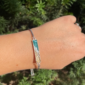 May include: A black adjustable bracelet with a silver bar charm. The bar charm is filled with green and gold glitter.