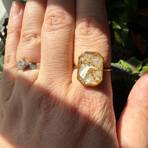 May include: A gold-toned ring with a rectangular, iridescent, white and gold stone. The ring has a thin band and a bezel setting. Another ring with a square diamond is also visible.