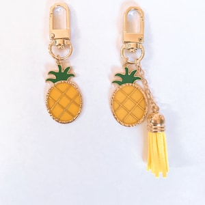 May include: Two gold-colored keychains with pineapple charms. The pineapple charms are yellow with green tops and gold outlines. One keychain has a yellow tassel attached.
