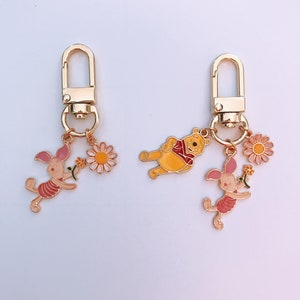 May include: Two gold keychains with charms. One keychain has a Winnie the Pooh charm and a pink piglet charm. The other keychain has a pink piglet charm and a yellow daisy charm.