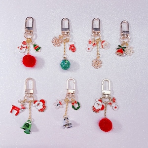 May include: A collection of Christmas-themed keychains. Each keychain features a gold-colored clasp and chain, with charms including snowmen, Christmas trees, snowflakes, Santa Claus, reindeer, bells, and a red pom-pom. The charms are in various colors.