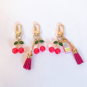 May include: Three gold cherry keychains with red glass cherries and green leaves. The keychain on the right has a multi-colored striped charm and a pink tassel.