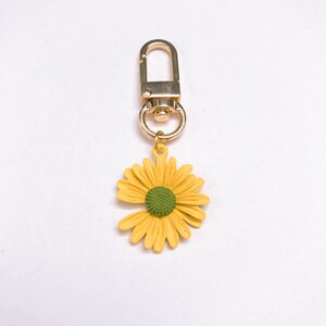 May include: A gold-colored keychain with a yellow daisy charm and a green center. The keychain has a lobster clasp for easy attachment. The daisy charm is a bright, cheerful accessory.