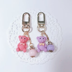 May include: Two gold keychains with pink and purple teddy bear charms and matching colored beads. The teddy bears have bows around their necks.