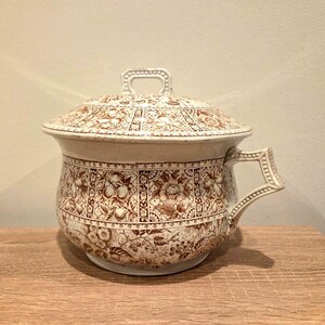 May include: A vintage ceramic chamber pot with a lid. The pot is decorated with a brown floral pattern on a cream background. It has a handle and a small handle on the lid. The pot is sitting on a wooden surface.