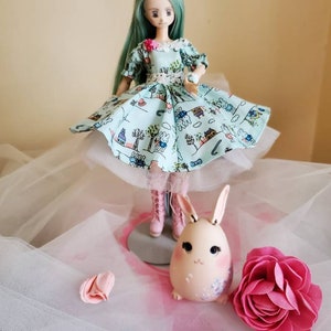 Può includere: Una bambola con lunghi capelli verde menta che indossa un vestito azzurro con una stampa stravagante di conigli e case. Una piccola statuina a forma di uovo di coniglio e un fiore di carta rosa sono anche nell'immagine.