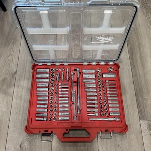 May include: A Milwaukee Packout tool set featuring a red plastic case with a clear lid. The interior contains a variety of silver metal sockets, extensions, and a ratchet wrench organised in moulded red slots. The Milwaukee logo is visible.