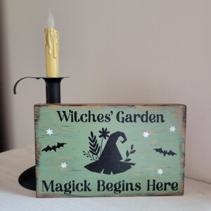 May include: A green wooden sign with the text "Witches' Garden Magick Begins Here" in black lettering. The sign features a black silhouette of a witch's hat with floral accents and bats.