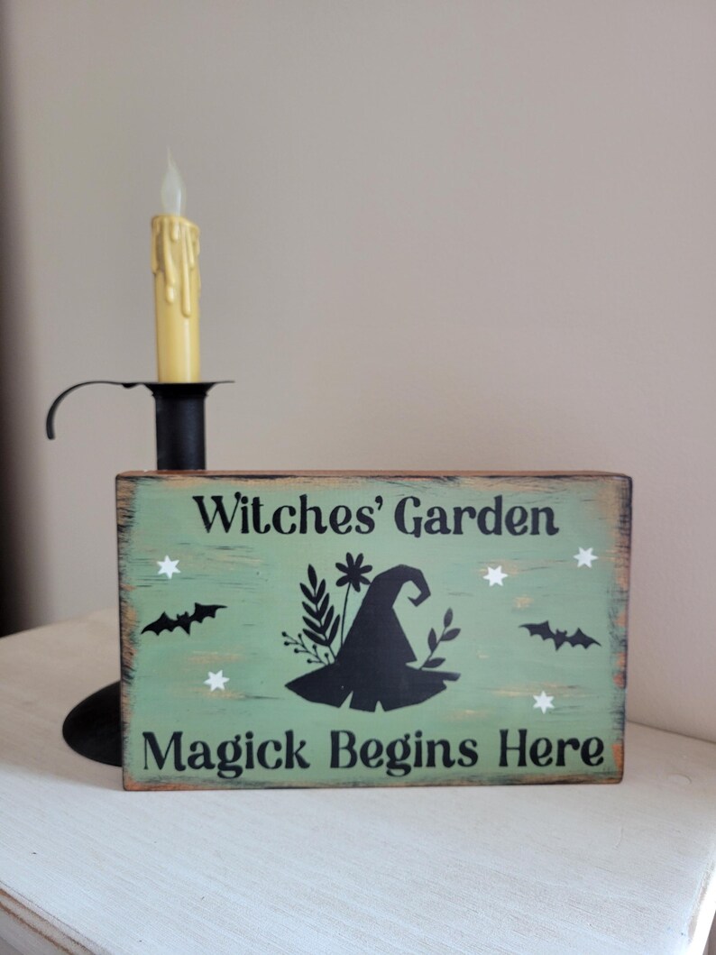 May include: A green wooden sign with the text "Witches' Garden Magick Begins Here" in black lettering. The sign features a black silhouette of a witch's hat with floral accents and bats.