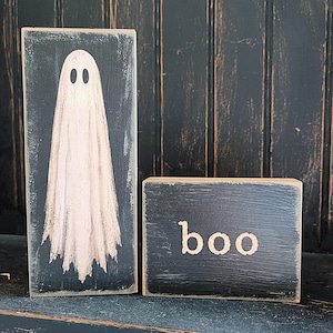 May include: Two rectangular wooden blocks with a distressed black finish. The taller vertical block features a painted white ghost with two black oval eyes. The shorter horizontal block features the word "boo" painted in a light beige color.