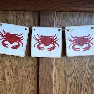 May include: A red crab garland with five white wooden squares, each featuring a red crab silhouette. The garland is strung with twine and hangs against a woodgrain background.