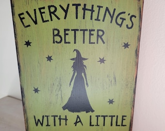 May include: A rectangular wooden sign with a distressed green background and black text that reads "EVERYTHING'S BETTER WITH A LITTLE MAGIC." A silhouette of a witch and stars are also featured on the sign.