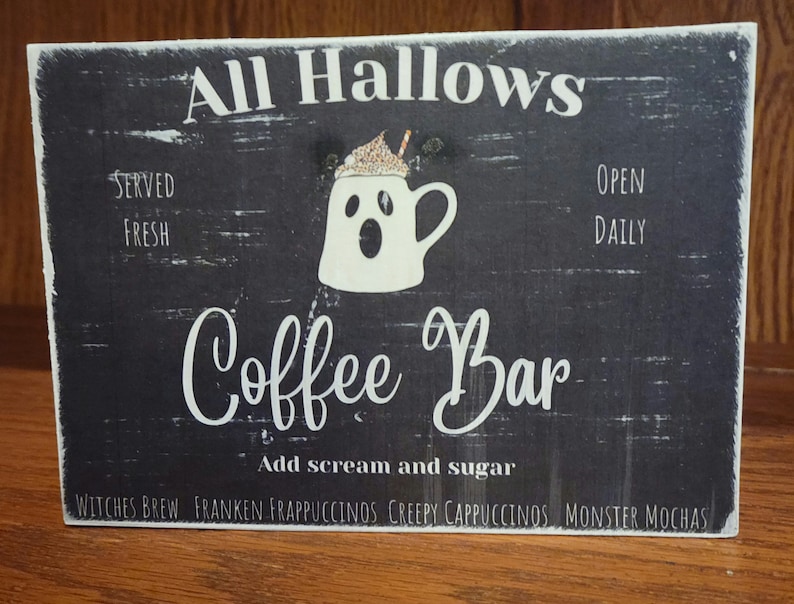 May include: A black and white sign with the text "All Hallows Coffee Bar" and a ghost-shaped coffee mug with whipped cream and a straw. The sign also says "Served Fresh" and "Open Daily". Below the main text, it says "Add scream and sugar" and lists "Witches Brew, Franken Frappuccinos, Creepy Cappuccinos, Monster Mochas".