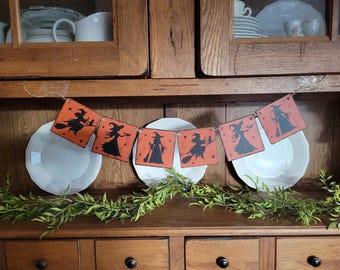 May include: A Halloween-themed banner with orange square pendants featuring black witch silhouettes. The banner is strung on twine and displayed in front of white plates and a wooden cabinet. Greenery adds a decorative touch.