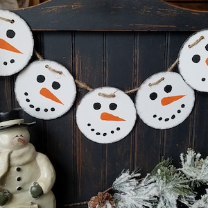 May include: A white snowman garland with five round ornaments featuring black eyes, orange noses, and black dots for mouths. The garland is strung with twine and hangs on a dark wood background.