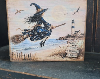 May include: A rectangular wooden sign featuring a witch in a blue star-patterned dress flying on a broomstick over a beach with a lighthouse in the background. The sign includes the text Salty Sand & Witchy Plans printed on a wooden signpost.
