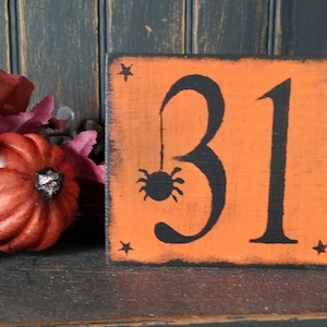 May include: A square orange wooden block with distressed black edges featuring the black number 31, a small spider hanging from the top of the number three, and four small black stars positioned in each corner of the block surface.
