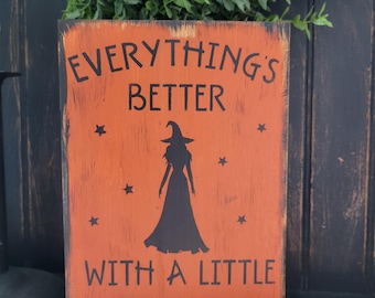 May include: Orange wooden sign with black text that reads "EVERYTHING'S BETTER WITH A LITTLE MAGIC." A black silhouette of a witch is in the center, surrounded by black stars. The sign has a distressed, rustic look.