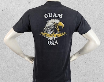 May include: A black short-sleeve t-shirt featuring a graphic on the back of a bald eagle head with a yellow beak. Below the eagle is a yellow banner with the text AMERICAN BIKER. The word GUAM is printed above the eagle and USA is printed below.