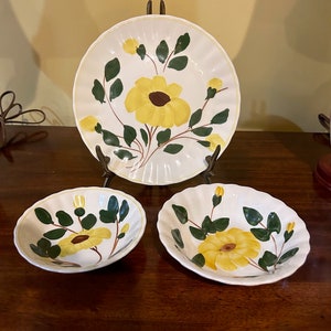 May include: A set of three white ceramic dishes with a yellow floral design. The set includes a large plate and two smaller bowls. The flowers are yellow with brown centers and green leaves. The edges of the plate and bowls are scalloped.