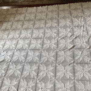 May include: A large, off-white crochet blanket featuring a repeating square pattern. Each square displays a central star or snowflake motif created with intricate thread work. The textile is composed of multiple joined squares, forming a uniform grid.