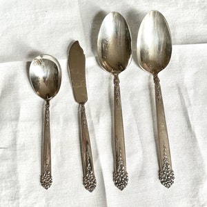 May include: Four pieces of vintage silverware are arranged on a white cloth. The set includes a small spoon, a butter knife, and two larger spoons. Each piece has a decorative floral design on the handle.