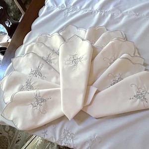 May include: A set of cream-colored fabric napkins arranged in a fan shape on a matching tablecloth. Each napkin features a scalloped edge and a delicate, light gray embroidered floral motif centered on the fabric surface.