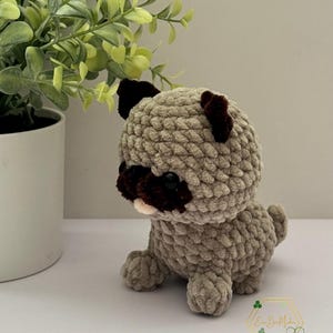 May include: A crocheted plush toy shaped like a pug dog. The toy is light brown with dark brown ears and a black nose. The toy is sitting on a white surface.
