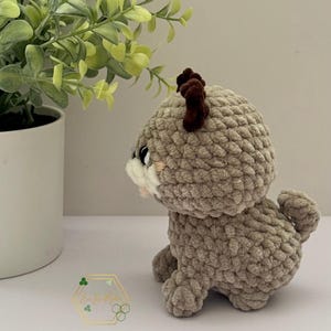 May include: A crocheted plush toy in the shape of a deer. The deer is light brown with a white face and brown antlers. The deer is sitting on a white surface.