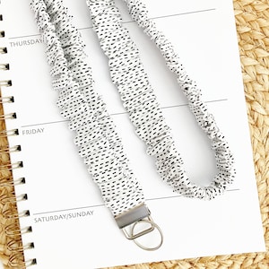 May include: A white lanyard with a black dotted pattern, featuring a silver-colored metal clasp and ring. The lanyard is lying on a white notebook with the days of the week printed on it.