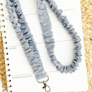 May include: A light blue fabric lanyard with a silver-colored clasp. The lanyard is made of gathered fabric and rests on a white notebook with gold spiral binding. The notebook is open to a page with the days of the week printed on it.