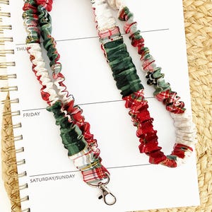 May include: A fabric lanyard featuring a gathered, scrunchie-style design with a festive plaid pattern in red, dark green, and white. The lanyard includes a silver-toned metal swivel lobster clasp at the bottom for attaching items securely.