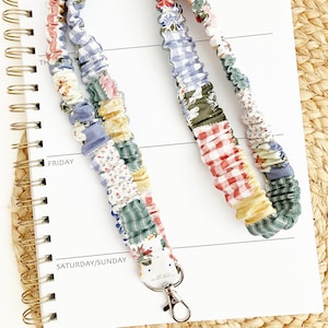 May include: A fabric lanyard featuring a gathered, scrunchie-style design made from a patchwork of various floral, gingham, and solid-colored cotton fabrics in shades of red, blue, yellow, green, and white, finished with a silver metal swivel clasp.