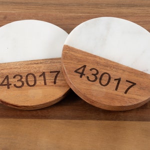 May include: Two round coasters with a white marble top and a wooden bottom. The wooden section is engraved with the number "43017". The coasters are set on a wooden surface.