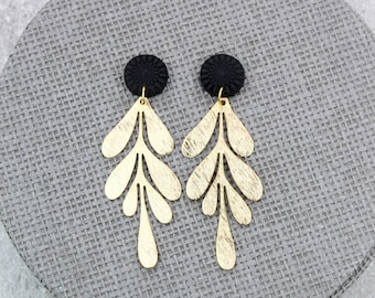 Puede incluir: Un par de pendientes dorados en forma de hoja con tachuelas circulares negras. Los pendientes tienen un acabado texturizado y se exhiben sobre una superficie gris texturizada. Joyas.